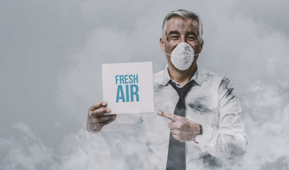 Man with a mask on, standing in a room full of smoke, and pointing at a sign that says "Fresh Air."