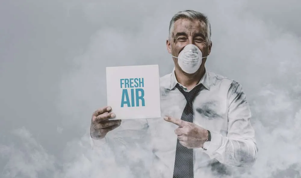 Man with a mask on, standing in a room full of smoke, and pointing at a sign that says "Fresh Air."