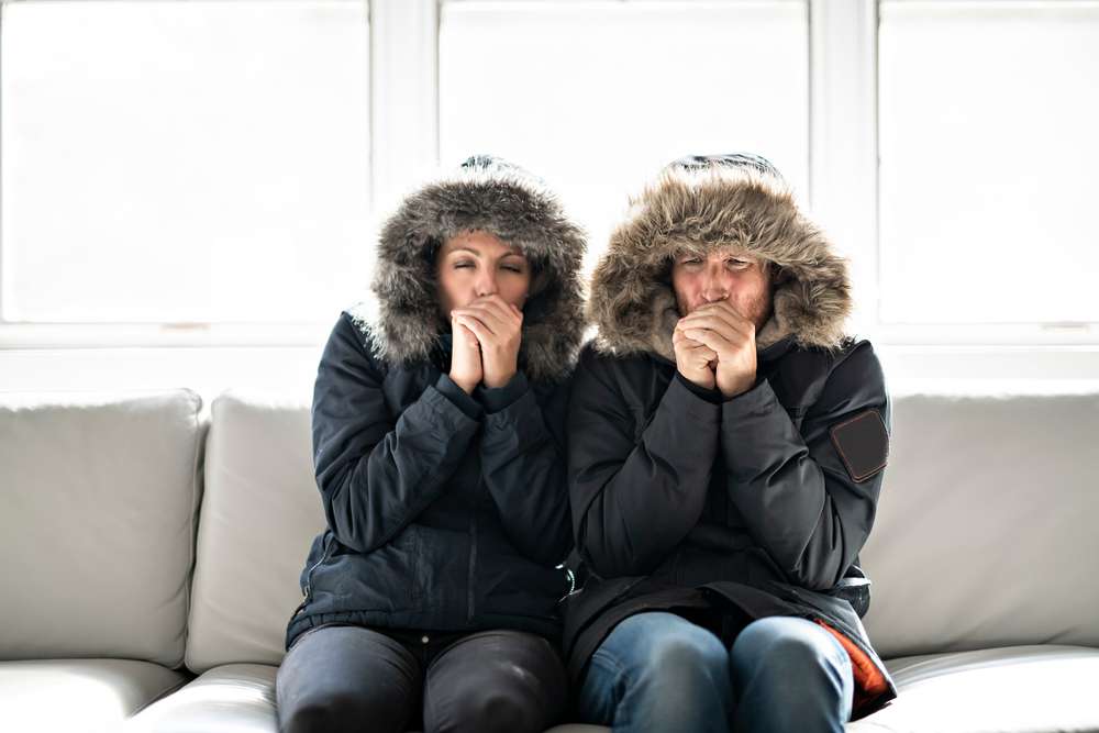 Couple sitting on their couch, dressed in winter coats, and blowing into their cupped hands to get warm.