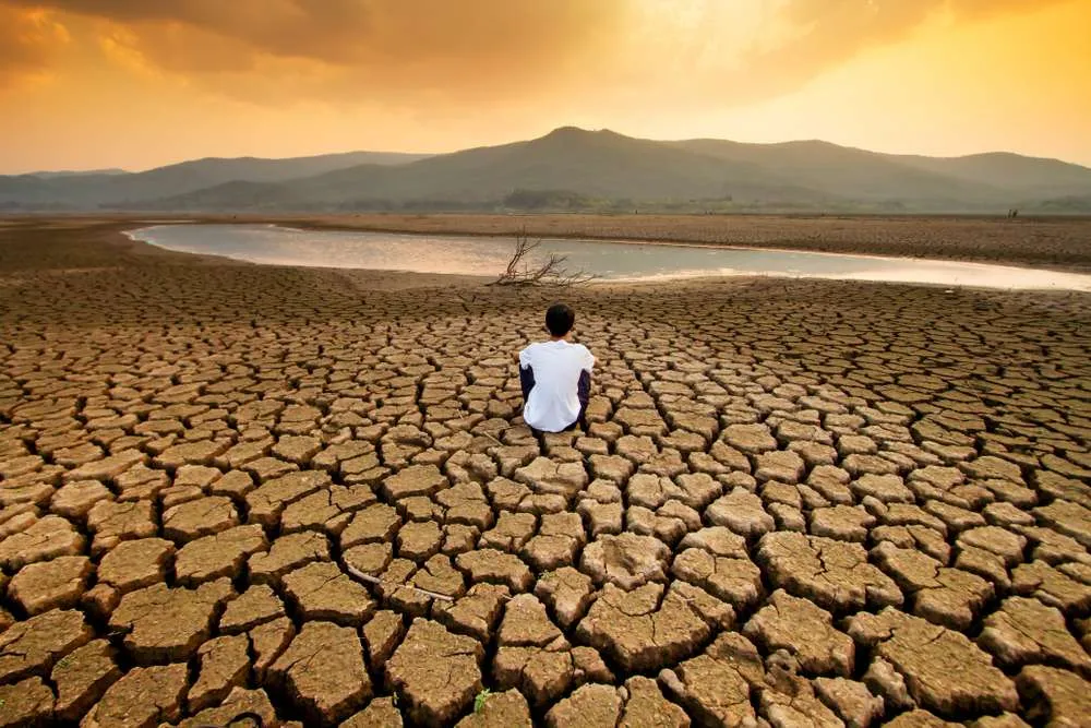 Man sitting on dry, cracked desert, staring at a lake.