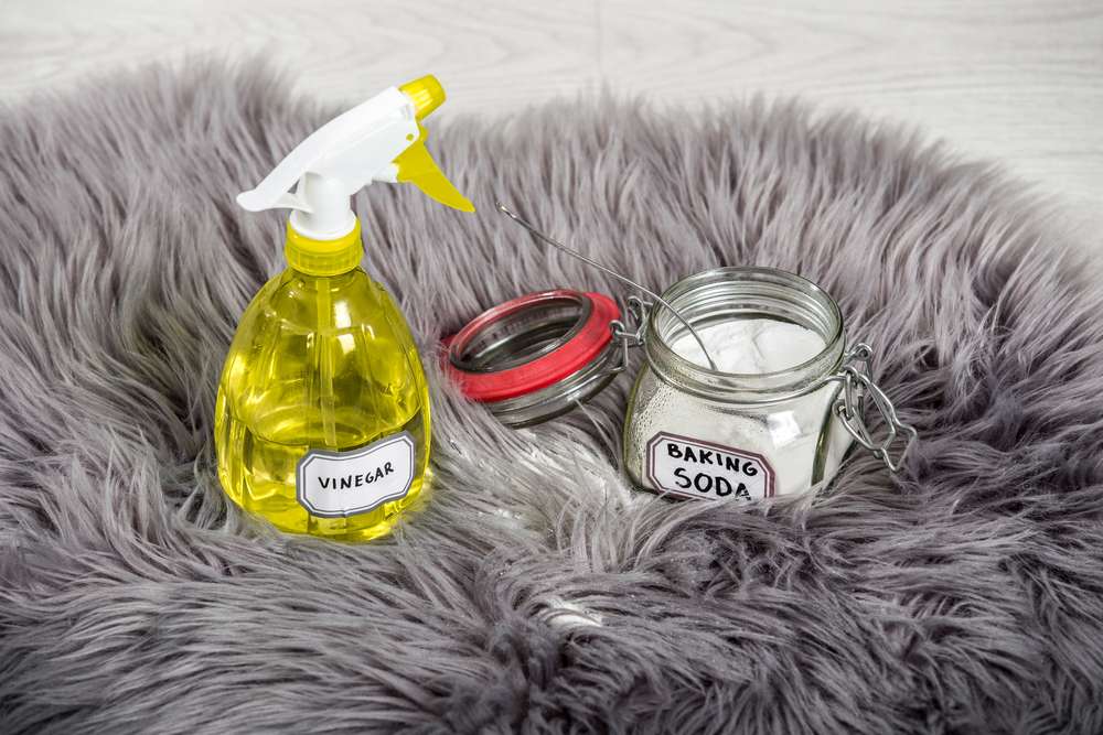 Spray bottle with vinegar in it sitting on a rug next to a jar of baking soda.